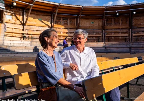 Coppia sorridente seduta su panchine di legno, con la Berlin WelcomeCard e il bus hop-on hop-off