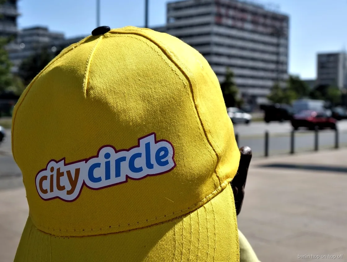 Primer pla d'una gorra groga City Circle amb logotip en un carrer assolellat, bus hop on hop off de Berlín