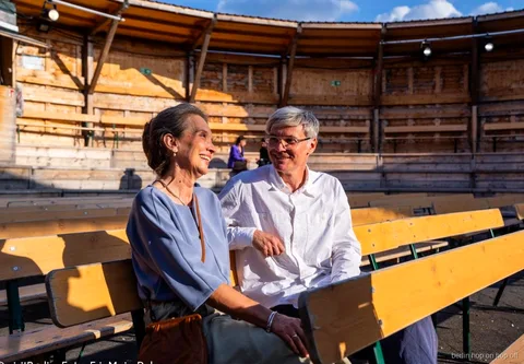 Smiling couple sitting on wooden benches, with the Berlin WelcomeCard and hop-on hop-off bus